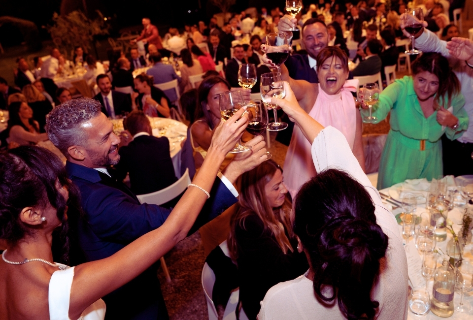 Un gruppo di persone in abiti formali alza i bicchieri in un brindisi celebrativo durante un grande e affollato banchetto.